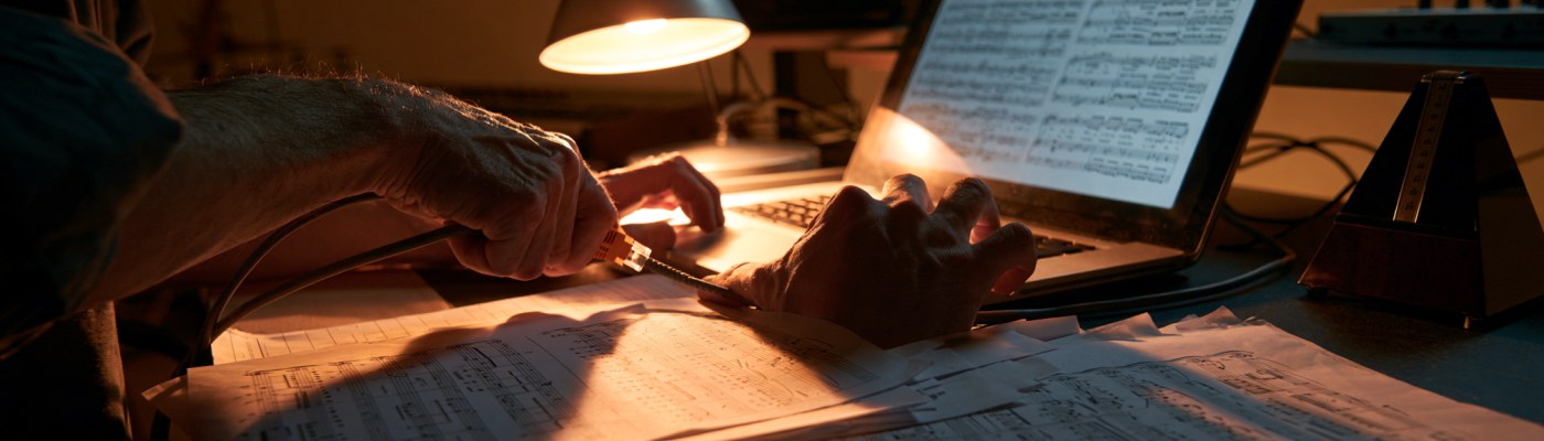 Hands connecting an ethernet cable to a laptop on a music studio desk, with sheet music and a metronome