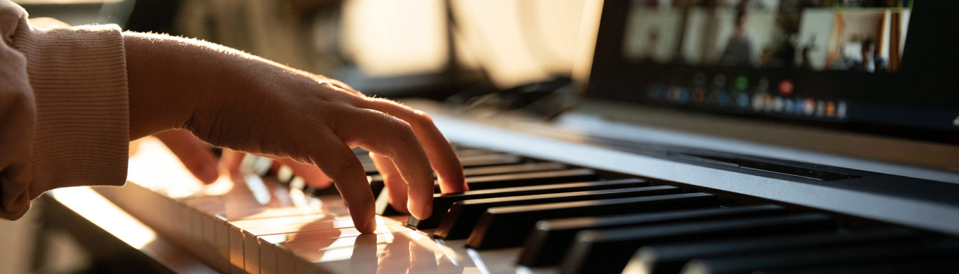 A child's hands on the piano keys during an online lesson, with a blurred laptop in the background