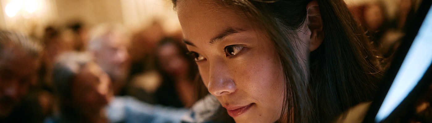 Close-up of a young pianist looking at a screen during a masterclass, with a blurred audience in the background