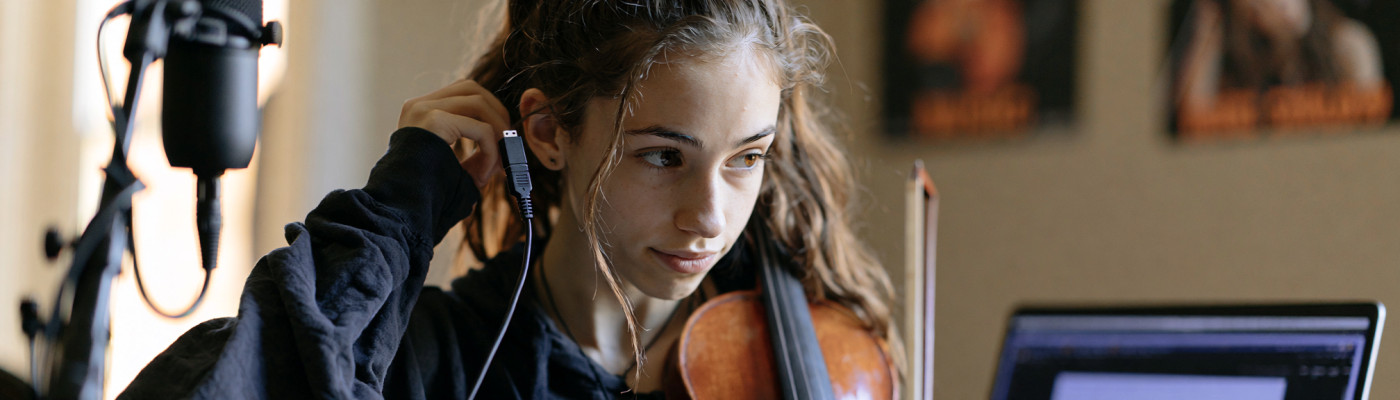 A teenager adjusting a USB microphone before an online music lesson, with a violin and bow resting on the desk next to the laptop
